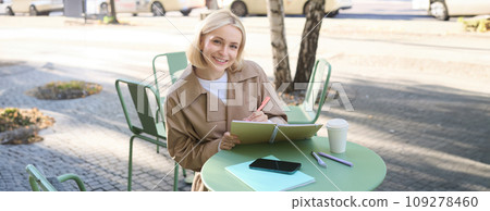 Portrait of beautiful blond woman, sitting in outdoor coffee shop, drawing in cafe in notebook, making sketches outside on street 109278460
