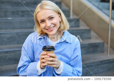 Close up portrait of female model, cute student with cup of coffee, sitting on stairs near campus, spending time outside 109278500