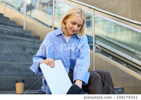 Portrait of young street-style girl, student with backpack, putting away laptop, packing, sitting on stairs with cup of coffee, wearing blue shirt 109278501