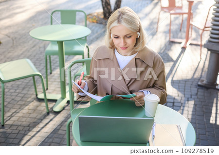 Portrait of young blond woman, female student in street cafe, wearing wireless headphones, using laptop, having online meeting, attend web lecture or course, doing homework 109278519