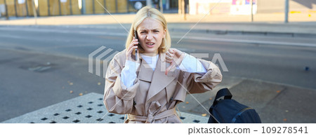 Disappointed young woman sitting on bench and showing thumbs down, talking on mobile phone and frowning, dislike conversation Disappointed young woman sitting on bench and showing thumbs down, talking on mobile phone and frowning, dislike conversation 109278541