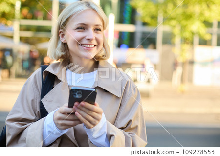 Carefree smiling woman standing on sunny street, using mobile phone, waiting for car or bus with smartphone, looking around 109278553