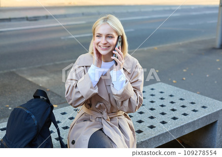 Image of young blond woman sitting on street bench with backpack, talking on mobile phone, answers a call and smiling while chatting with someone Image of young blond woman sitting on street bench with backpack, talking on mobile phone, answers a call and smiling while chatting with someone 109278554