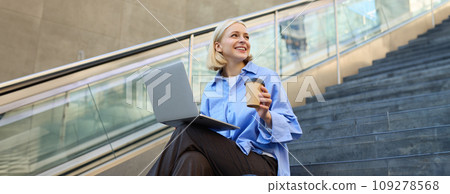 Young woman, student in blue shirt, working on laptop, sitting outdoors on street stairs, working on project online, connects to public wifi, elearning 109278568