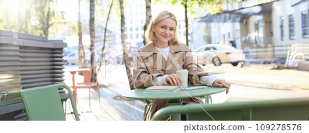 Portrait of young blonde woman sitting in cafe, doing homework and drinking coffee, writiing in journal, using notebook on fresh air 109278576