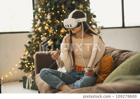 In the snug setting of a Christmas celebration at home, a fashionable young lady utilizes a virtual reality headset. In the snug setting of a Christmas celebration at home, a fashionable young lady utilizes a virtual reality headset. 109278600