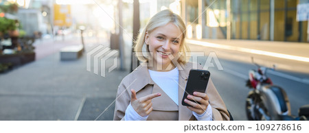 Close up portrait of young blond woman, student pointing at smartphone, standing on street and showing smth on mobile phone 109278616
