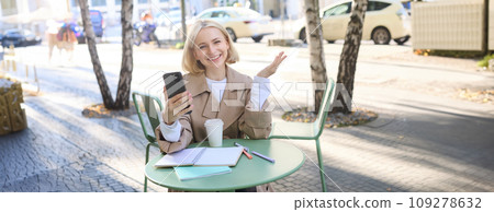 Happy young blond female model in coffee shop, talks on video chat, using smartphone and having conversation while resting outdoors in city centre Happy young blond female model in coffee shop, talks on video chat, using smartphone and having conversation while resting outdoors in city centre 109278632