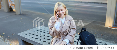 Image of carefree young candid woman, sitting on bench outside, talking on mobile phone, having relaxed unbothered face during conversation 109278633