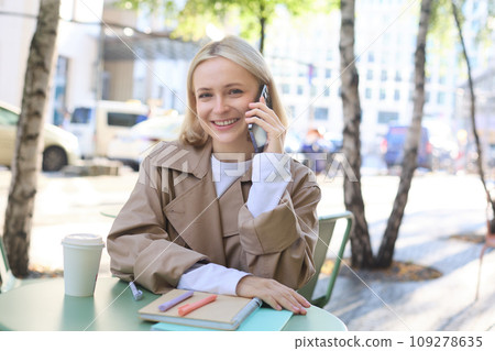 Close up of cheerful blond girl, woman talking on mobile phone, sitting in city centre, outdoor cafe, taking break from work or studying, chatting on telephone Close up of cheerful blond girl, woman talking on mobile phone, sitting in city centre, outdoor cafe, taking break from work or studying, chatting on telephone 109278635