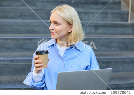 Young woman sitting with cup of coffee and laptop on stairs outdoors, working remotely, freelancing, connecting to public wifi in city to study online 109278641