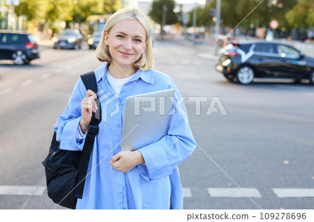 Street stylish portrait of young happy woman, blond girl with backpack and laptop, standing outdoor s near road on sunny bright day 109278696