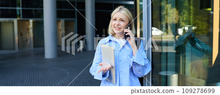 Close up portrait of modern young woman, student standing near building in city centre, talking on mobile phone, having chat on smartphone with confused face expression Close up portrait of modern young woman, student standing near building in city centre, talking on mobile phone, having chat on smartphone with confused face expression 109278699