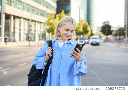Portrait of young city girl, urban woman with backpack, reading messages on smartphone and smiling, checking notifications, standing on street Portrait of young city girl, urban woman with backpack, reading messages on smartphone and smiling, checking notifications, standing on street 109278723