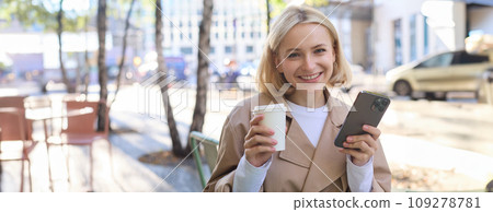 Portrait of cute blond woman with smartphone, drinking chai, coffee at favourite place in city centre, sitting outdoors, holding mobile phone Portrait of cute blond woman with smartphone, drinking chai, coffee at favourite place in city centre, sitting outdoors, holding mobile phone 109278781