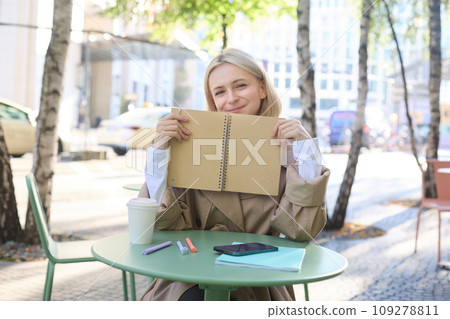 Portrait of young woman sitting in outdoor cafe, hiding behind journal, holding notebook in hands and smiling Portrait of young woman sitting in outdoor cafe, hiding behind journal, holding notebook in hands and smiling 109278811