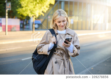 Image of young modern woman, student waiting for someone on street, standing with backpack and checking her mobile phone, looking at smartphone screen 109278856