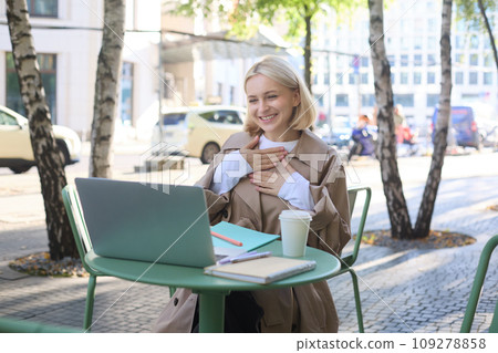 Image of young smiling woman working remotely, connects to online call, chatting via laptop, looking pleased, feeling grateful, laughing, sitting in outdoor cafe 109278858