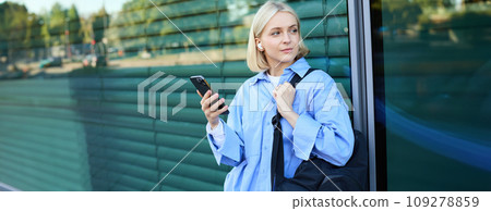 Lifestyle portrait of smiling young female model, student with backpack, waiting for someone on street, standing outdoors with smartphone 109278859