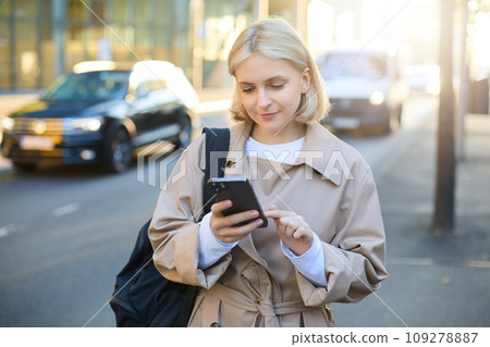 Close up portrait of young modern woman on street, using mobile phone, looking at smartphone screen, reading notification, waiting for taxi near road 109278887