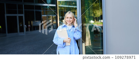 Lifestyle portrait of young woman with laptop, posing near campus, entrance to office building, holding backpack, smiling and looking happy 109278971