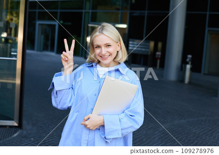 Young positive female model, student with notebooks and study material showing peace sign, standing near university, college campus, promo for new education scheme 109278976