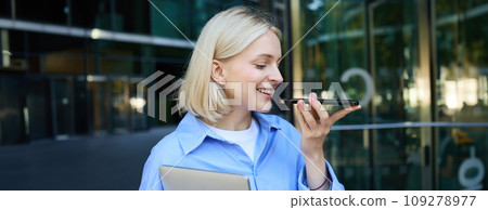 Close up portrait of smiling young female student, woman records voice message on smartphone, speaking on speakerphone while standing near office building, holding laptop Close up portrait of smiling young female student, woman records voice message on smartphone, speaking on speakerphone while standing near office building, holding laptop 109278977