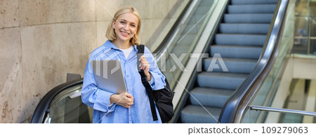Image of young modern woman, student on escalator, holding laptop and backpack, walking in city Image of young modern woman, student on escalator, holding laptop and backpack, walking in city 109279063
