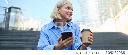 Lifestyle picture of smiling, beautiful young woman, drinking coffee, using mobile phone, resting in city, sitting on stairs 109279088