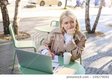 Lifestyle and technology concept. Young blond working woman in trench coat, studying, working on project on laptop, sitting in coffee shop on sunny day, making notes, attending online course 109279092