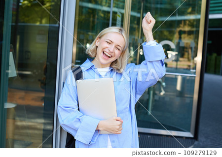 Carefree blond woman laughing, smiling and celebrating, posing with laptop near office, campus building, raising hand up in triumph, feeling excited Carefree blond woman laughing, smiling and celebrating, posing with laptop near office, campus building, raising hand up in triumph, feeling excited 109279129