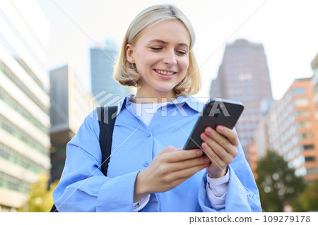 Close up portrait of stylish young blonde woman, standing on street, checking her mobile phone, using smartphone app to get around town, looking at online map for directions Close up portrait of stylish young blonde woman, standing on street, checking her mobile phone, using smartphone app to get around town, looking at online map for directions 109279178
