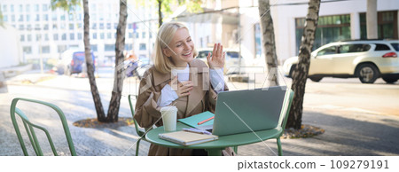 Portrait of young friendly woman, waving hand at laptop and smiling, saying hello, greeting someone, online chatting, having remote conversation while sitting in coffee shop Portrait of young friendly woman, waving hand at laptop and smiling, saying hello, greeting someone, online chatting, having remote conversation while sitting in coffee shop 109279191
