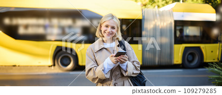 Street style shot of smiling young blond woman, holding mobile phone, standing on street and looking happy, holding backpack and wearing trench coat Street style shot of smiling young blond woman, holding mobile phone, standing on street and looking happy, holding backpack and wearing trench coat 109279285