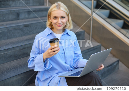 Portrait of blond smiling woman, drinking coffee, using laptop, sitting on campus stairs outside of building, studying, e-learning, connecting to wifi 109279310