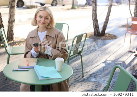 Portrait of smiling young woman using mobile phone while doing homework, working on project, sitting outdoors in cafe, drinking coffee and studying, holding smartphone 109279311