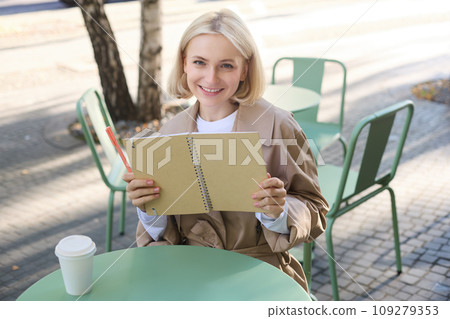 Portrait of young beautiful blond woman, artsy girl in coffee shop, holding notebook and pen, writing in her journal, drawing sketches outdoors 109279353