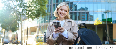 Lifestyle portrait of cute blonde girl with smartphone, sitting on bench with backpack, using mobile phone, reading message 109279462