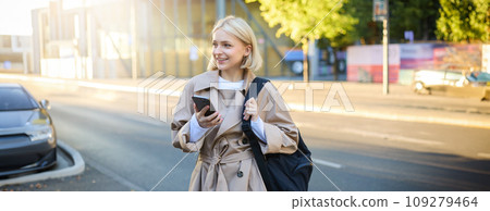 Modern young woman with backpack, student using smartphone, standing on street, sunny day, waiting for ride, using mobile phone application 109279464