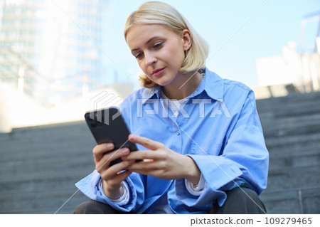 Close up portrait of young stylish female model, sitting outdoors on stairs, using mobile phone, reading on smartphone, messaging while waiting for someone outside 109279465