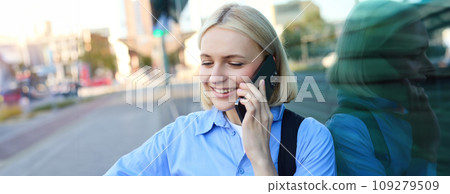 Close up portrait of blond smiling woman, answers a phone call, talking on telephone, standing on street and chatting, looking carefree and happy Close up portrait of blond smiling woman, answers a phone call, talking on telephone, standing on street and chatting, looking carefree and happy 109279509