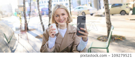 Beautiful young blond woman, posing with takeaway cup in outdoor cafe, sitting on street with coffee, taking selfie, recording live stream from favourite shop 109279521