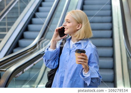 Portrait of cute young blond woman, student with cup of coffee, answers phone call, standing near escalator in city and talking, chatting with someone over the smartphone Portrait of cute young blond woman, student with cup of coffee, answers phone call, standing near escalator in city and talking, chatting with someone over the smartphone 109279534