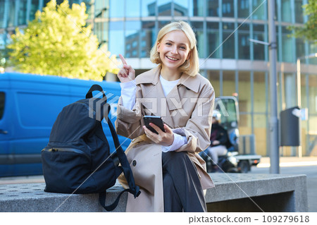 Portrait of young happy female student, woman sits on bench with backpack and phone, pointing finger at upper left corner, showing advertisement or banner 109279618