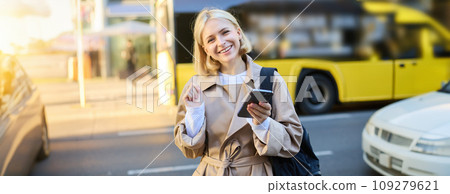 Street style shot of smiling young blond woman, holding mobile phone, standing on street and looking happy, holding backpack and wearing trench coat 109279621