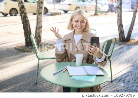Image of young woman working outdoors in cafe, sitting outside with smartphone, video chatting, connecting to online meeting using mobile phone, smiling and looking happy 109279799