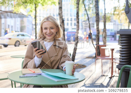 Portrait of blond smiling woman with smartphone, holding cup of coffee, drinking chai and enjoying sunny day outdoors in city centre 109279835