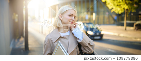 Portrait of smiling young woman with notebook, tucks hair behind her ear, walking along street, female model going to work, student coming home from university, carries notes and documents 109279880