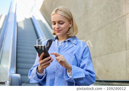 Close up portrait of young blond woman in city centre, standing near escalator, checking her mobile phone, reading message on smartphone Close up portrait of young blond woman in city centre, standing near escalator, checking her mobile phone, reading message on smartphone 109280012