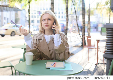 Portrait of woman, student doing homework outdoors in cafe, holding pen, frowning while calling someone, struggling to understand phone conversation Portrait of woman, student doing homework outdoors in cafe, holding pen, frowning while calling someone, struggling to understand phone conversation 109280014